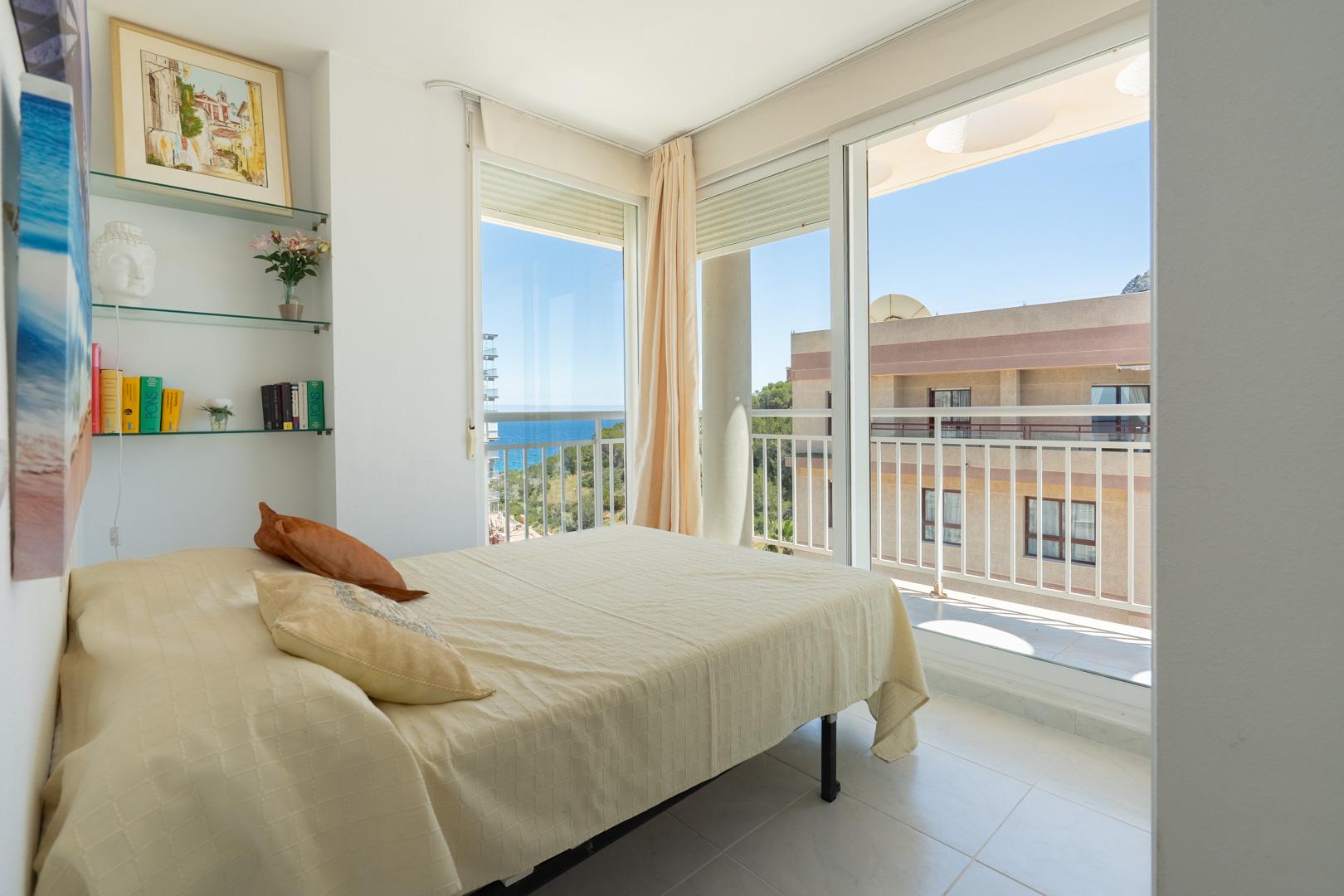 Helles Schlafzimmer mit Doppelbett, dekorativen Glasregalen und direktem Zugang zur Terrasse mit teilweisem Meerblick und Stadtansicht.