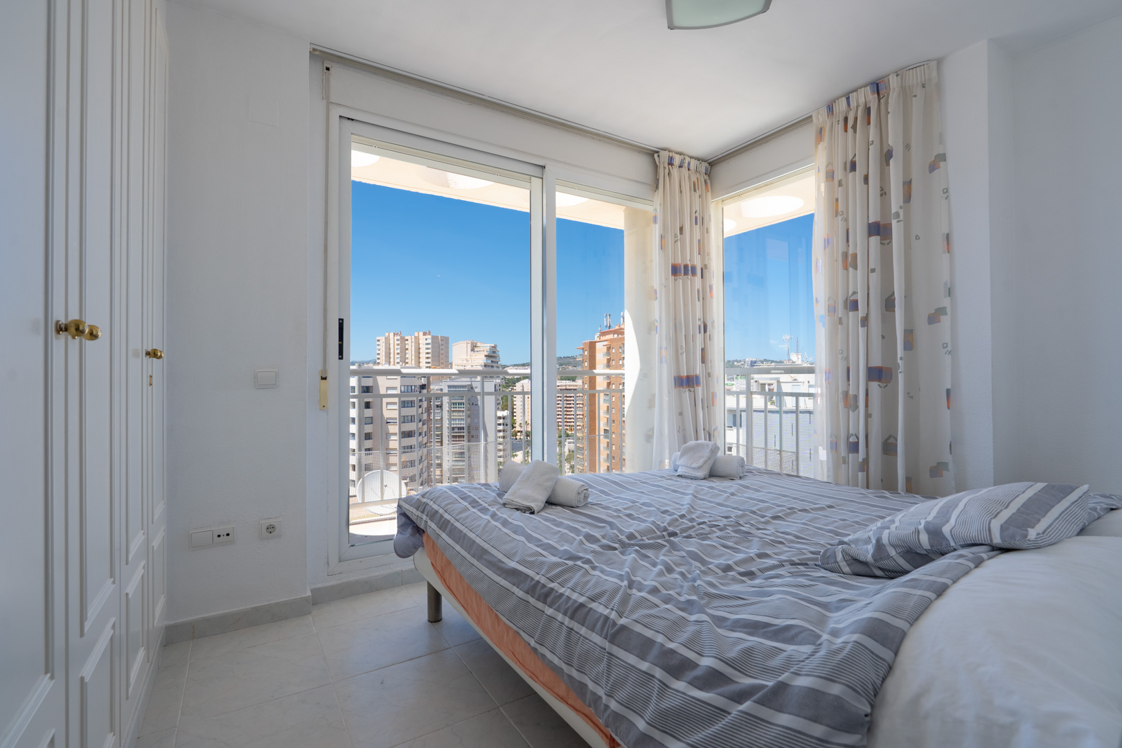 Helles Schlafzimmer mit Doppelbett, Einbauschrank und Glasschiebetüren zur Terrasse. Bietet klaren Stadtblick und blauen Himmel.