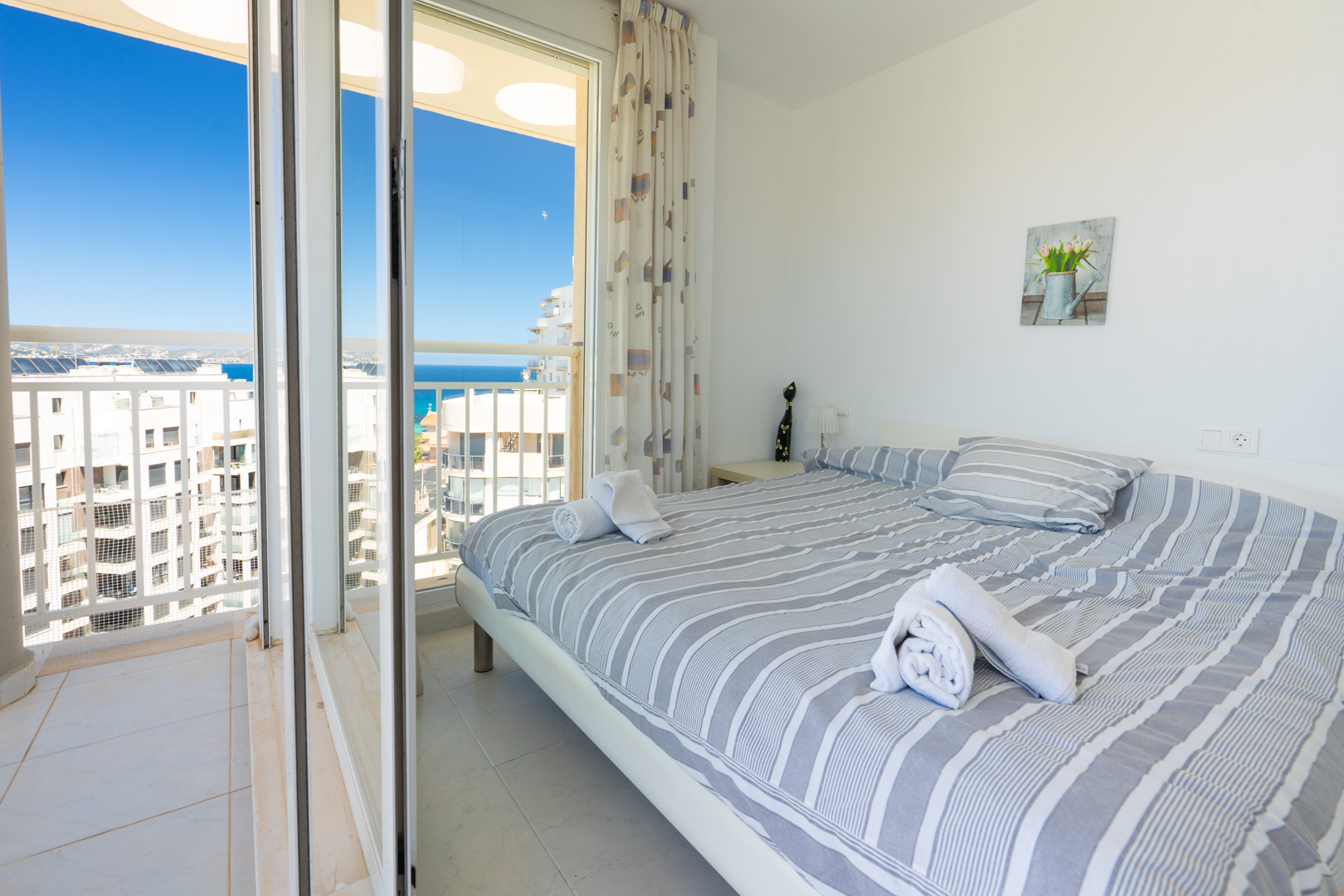 Helles Schlafzimmer mit Doppelbett, gestreifter Bettwäsche, Nachttischlampe und direktem Zugang zum privaten Balkon mit Meer- und Stadtblick.