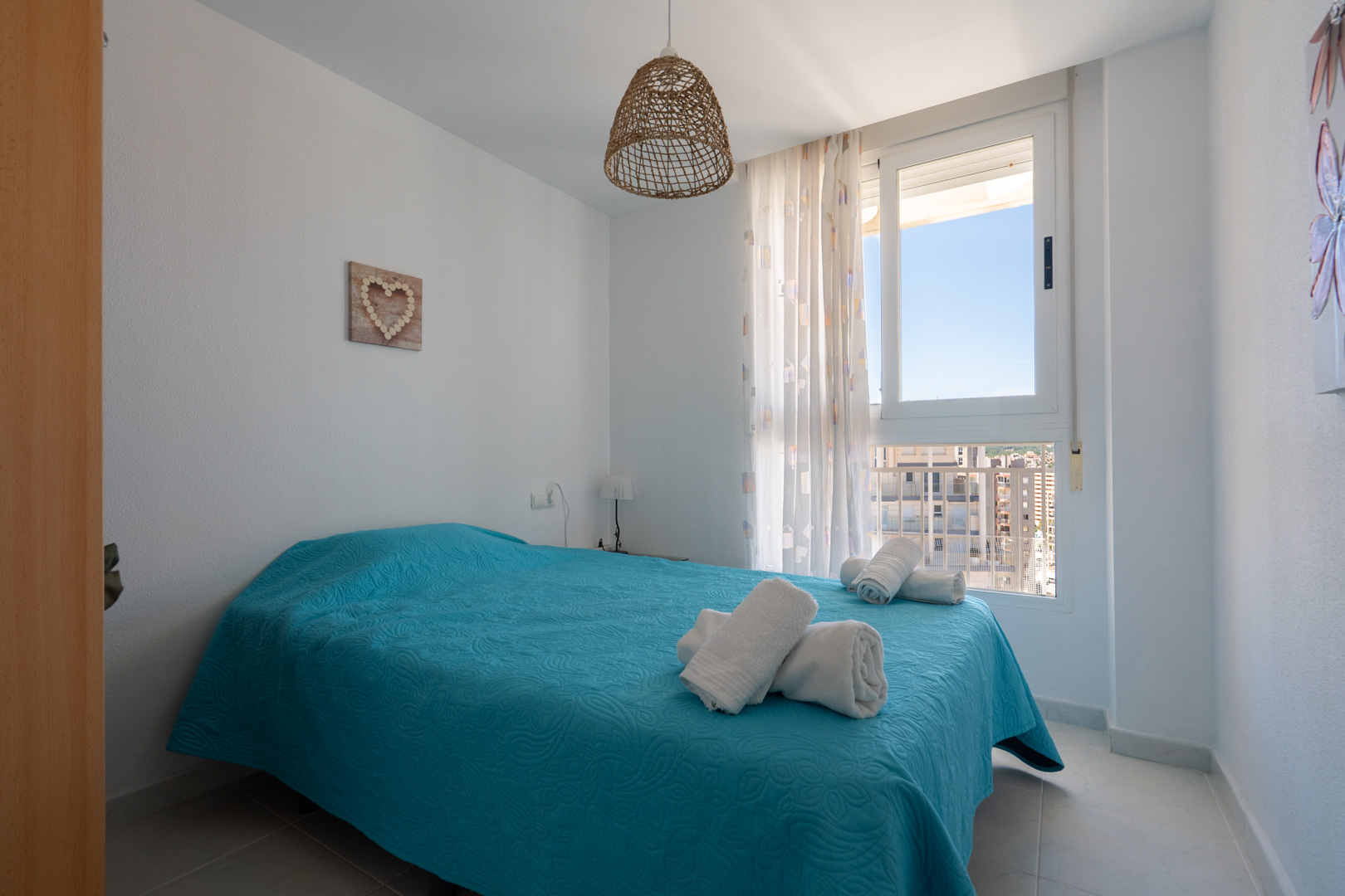 Helles Schlafzimmer mit Doppelbett in Türkis, einer Hängeleuchte aus Naturfasern und einem Fenster mit urbanem Blick auf umliegende Gebäude.