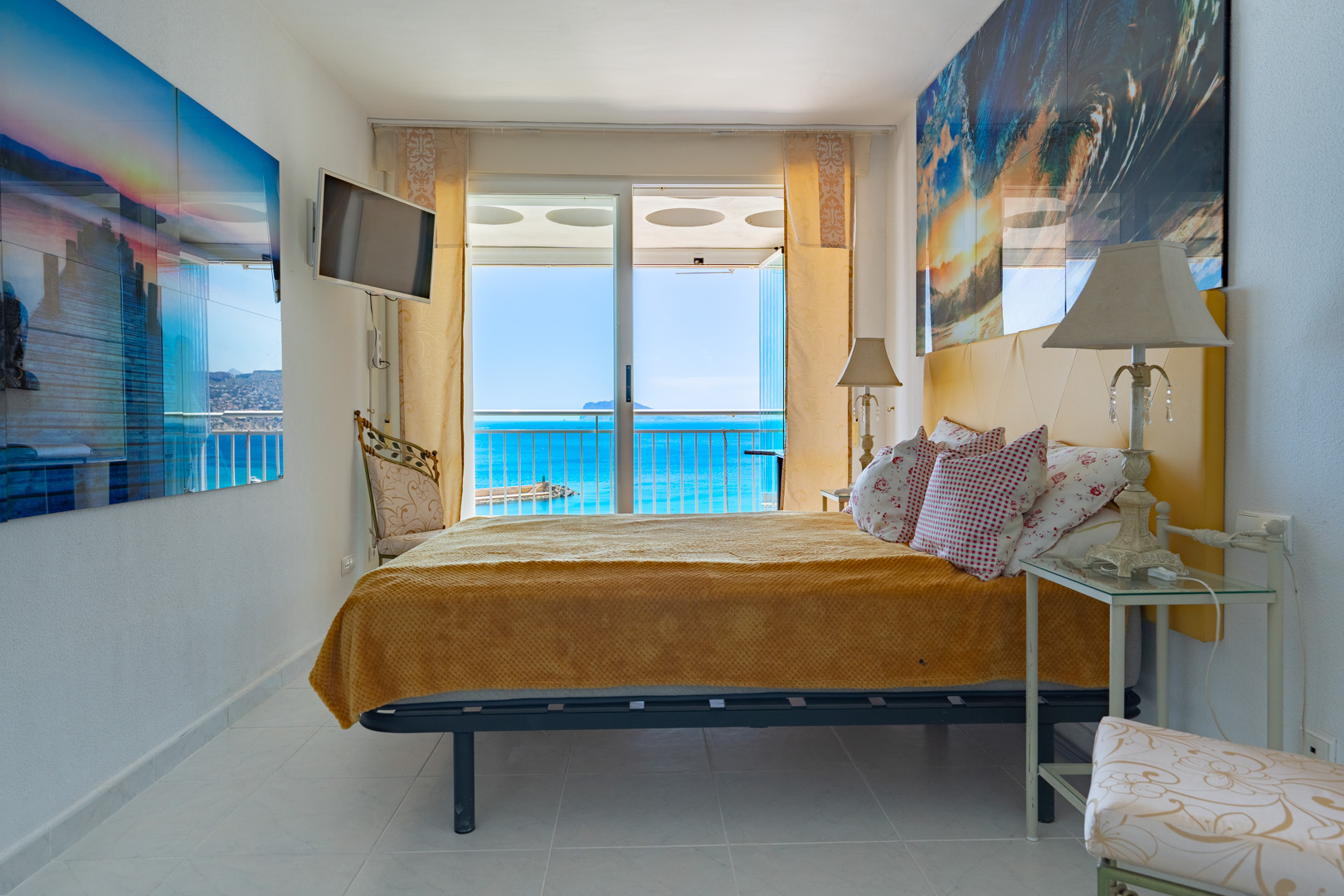 Helles Schlafzimmer mit Doppelbett und gelbem Kopfteil, weißen Fliesen und direktem Zugang zur Terrasse durch Schiebetüren, mit weitem Blick auf das Meer und den Hafen.