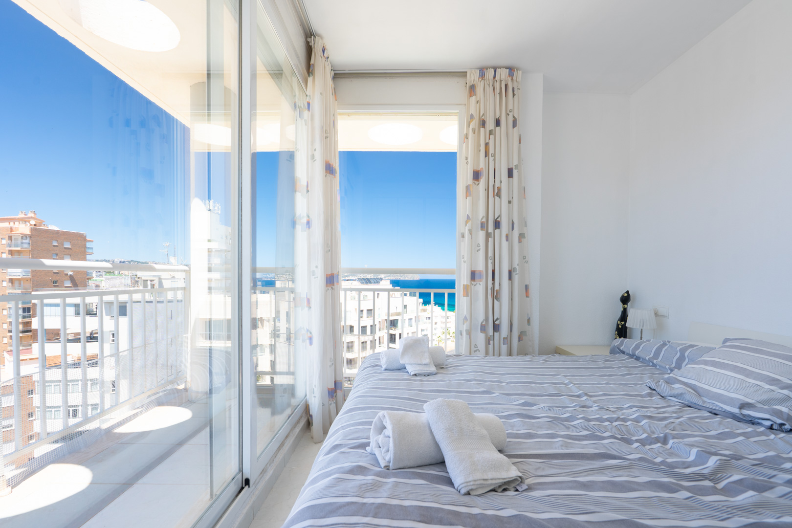 Helles Schlafzimmer mit Doppelbett und gestreifter Bettwäsche, direktem Zugang zur Terrasse und Panoramablick auf Meer und Himmel.