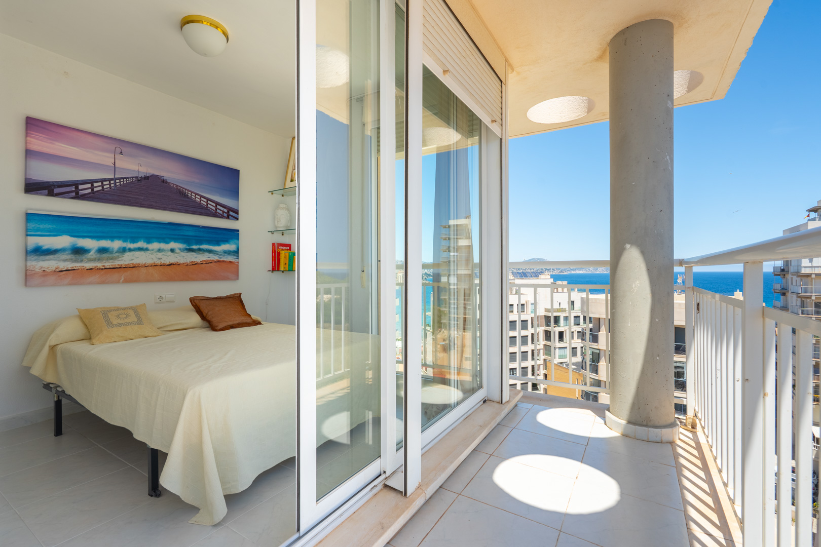 Helles Schlafzimmer mit Einzelbett und maritimer Wandkunst. Direkter Zugang zu einem privaten Balkon mit weißem Geländer, Betonsäule und Blick auf Meer und Küste.