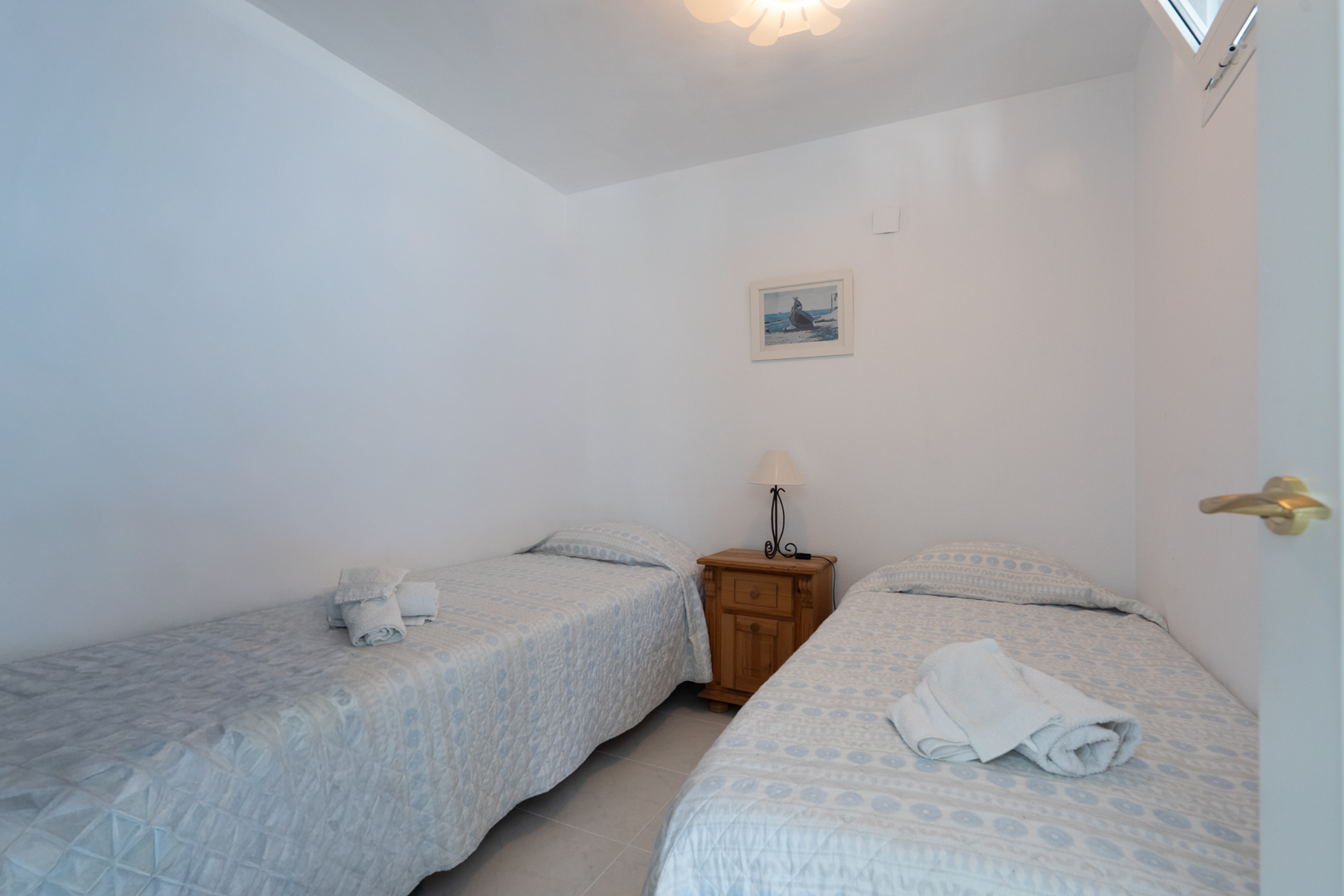 Helles Schlafzimmer mit zwei Einzelbetten, hell gemusterten Tagesdecken, einem Nachttisch aus Holz mit Lampe und einem kleinen Fenster.