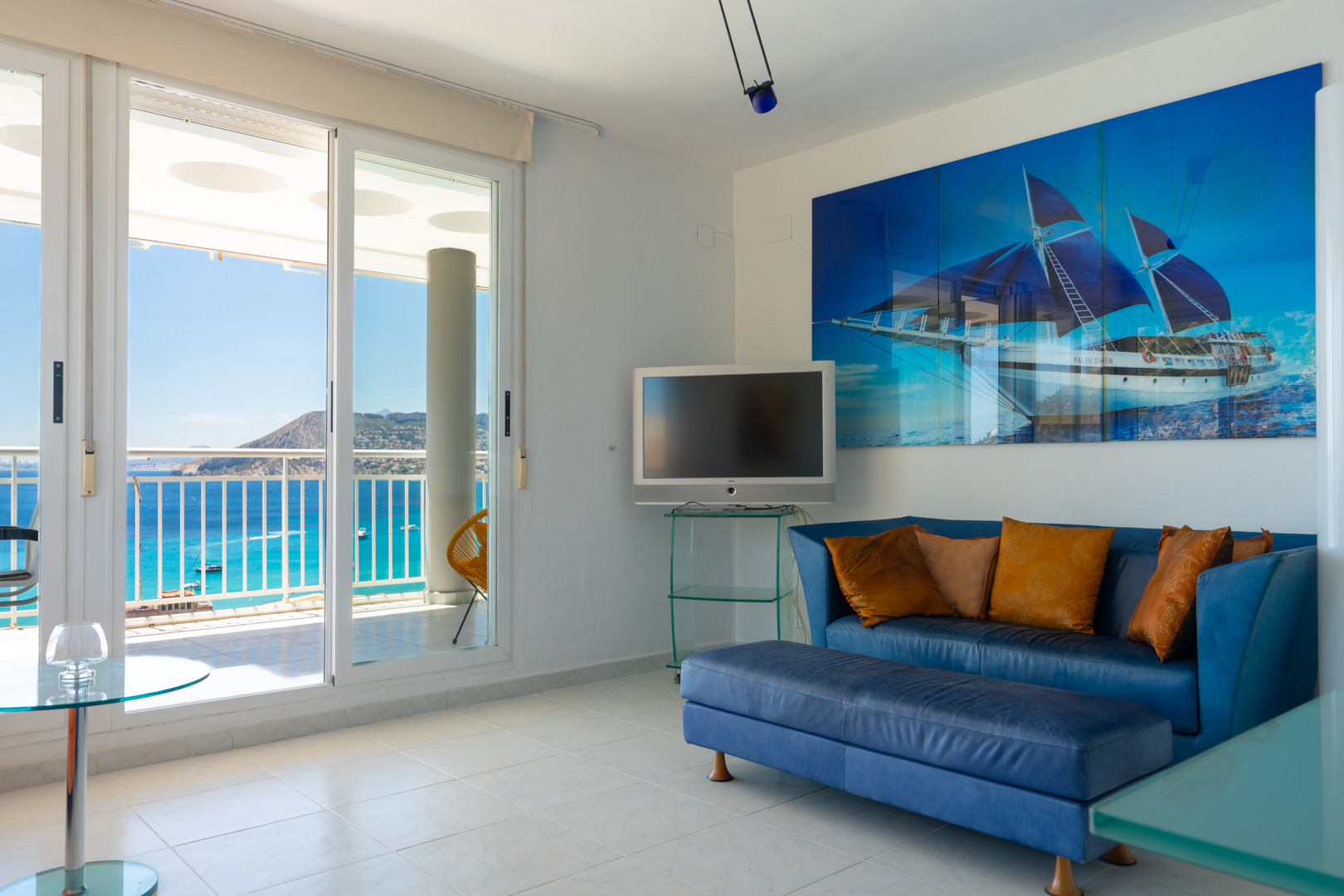 Helles Wohnzimmer mit Glasschiebetüren zur Terrasse und Panoramameerblick. Ausgestattet mit einem blauen Sofa, einem Wandfernseher und einem großen Bootsgemälde.