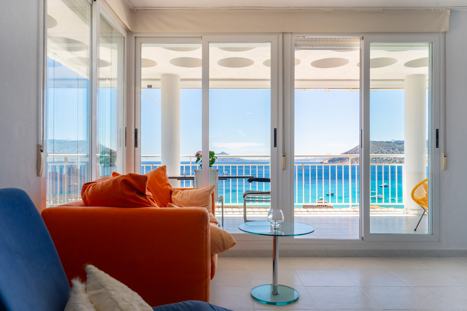 Helles Wohnzimmer mit orangefarbenem Sofa und Glastisch. Große Schiebetüren öffnen zu einer Terrasse mit direktem Meerblick auf Boote und ferne Berge.