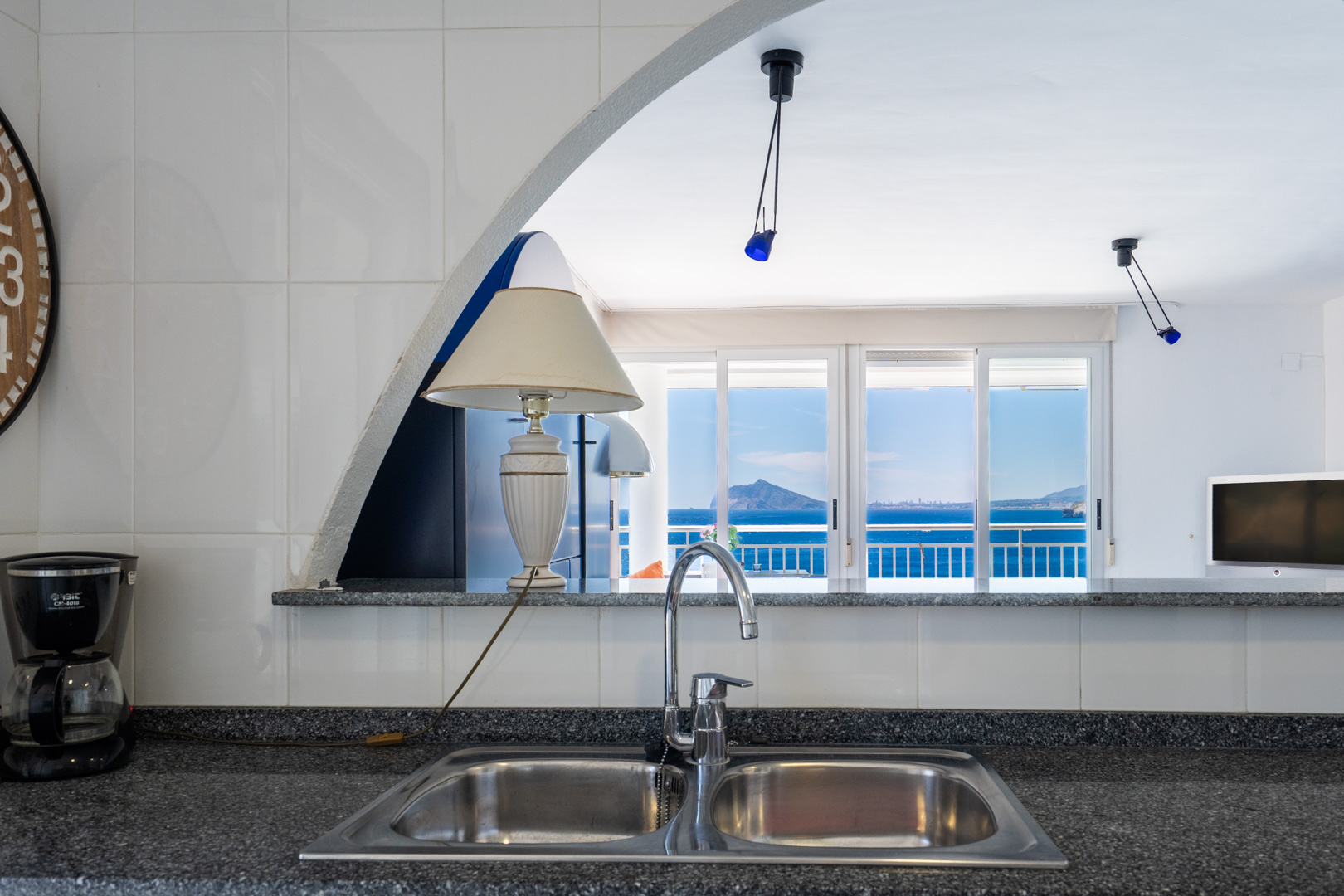 Küchenbereich mit Doppelspüle und dunkler Arbeitsplatte, durch einen Bogen mit dem Wohnbereich verbunden, der eine Terrasse mit weitem Meerblick freigibt.
