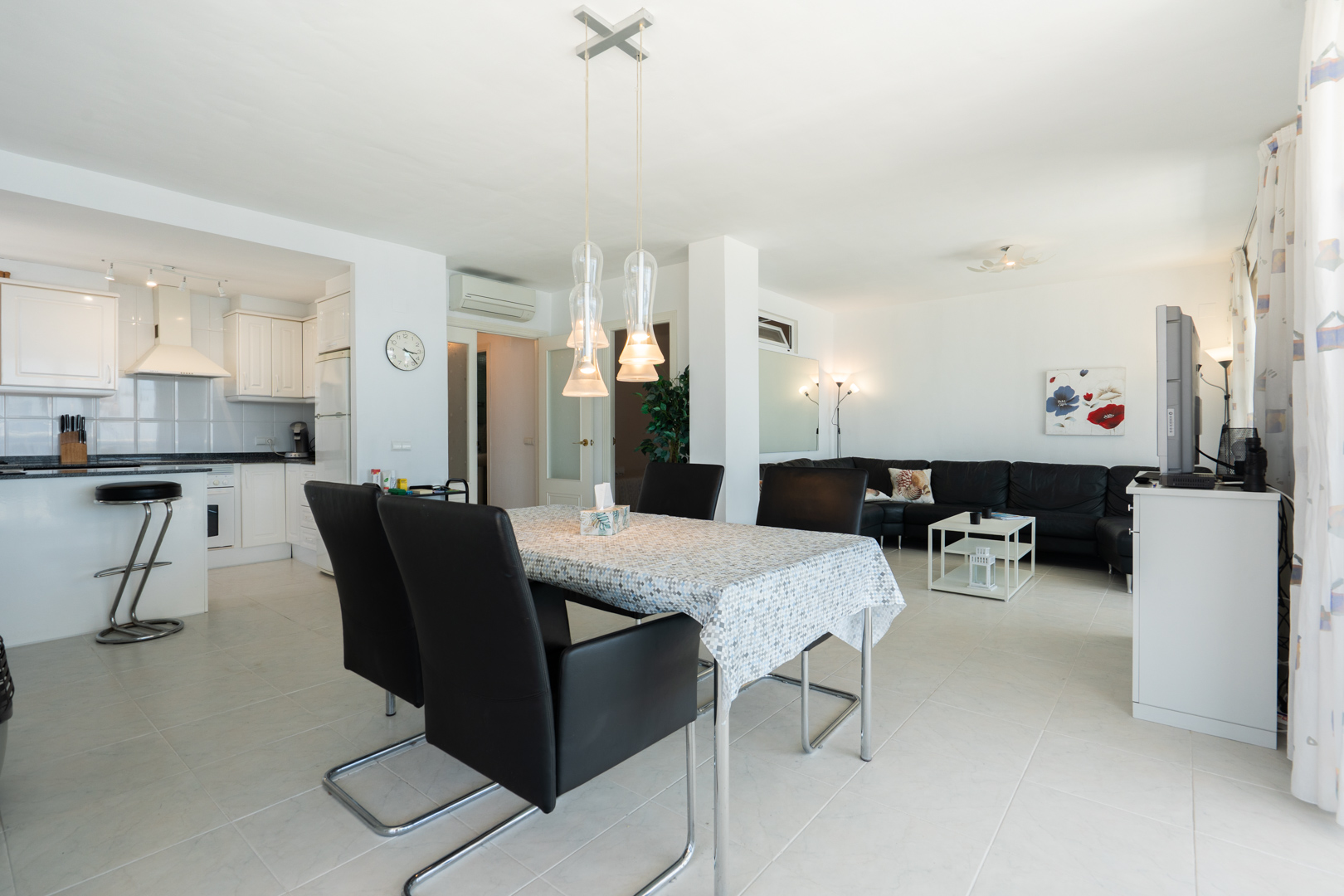 Offener Wohnbereich mit weißer Küche, Frühstücksbar, Essbereich mit moderner Pendelleuchte und Wohnzimmer mit schwarzem Ecksofa.