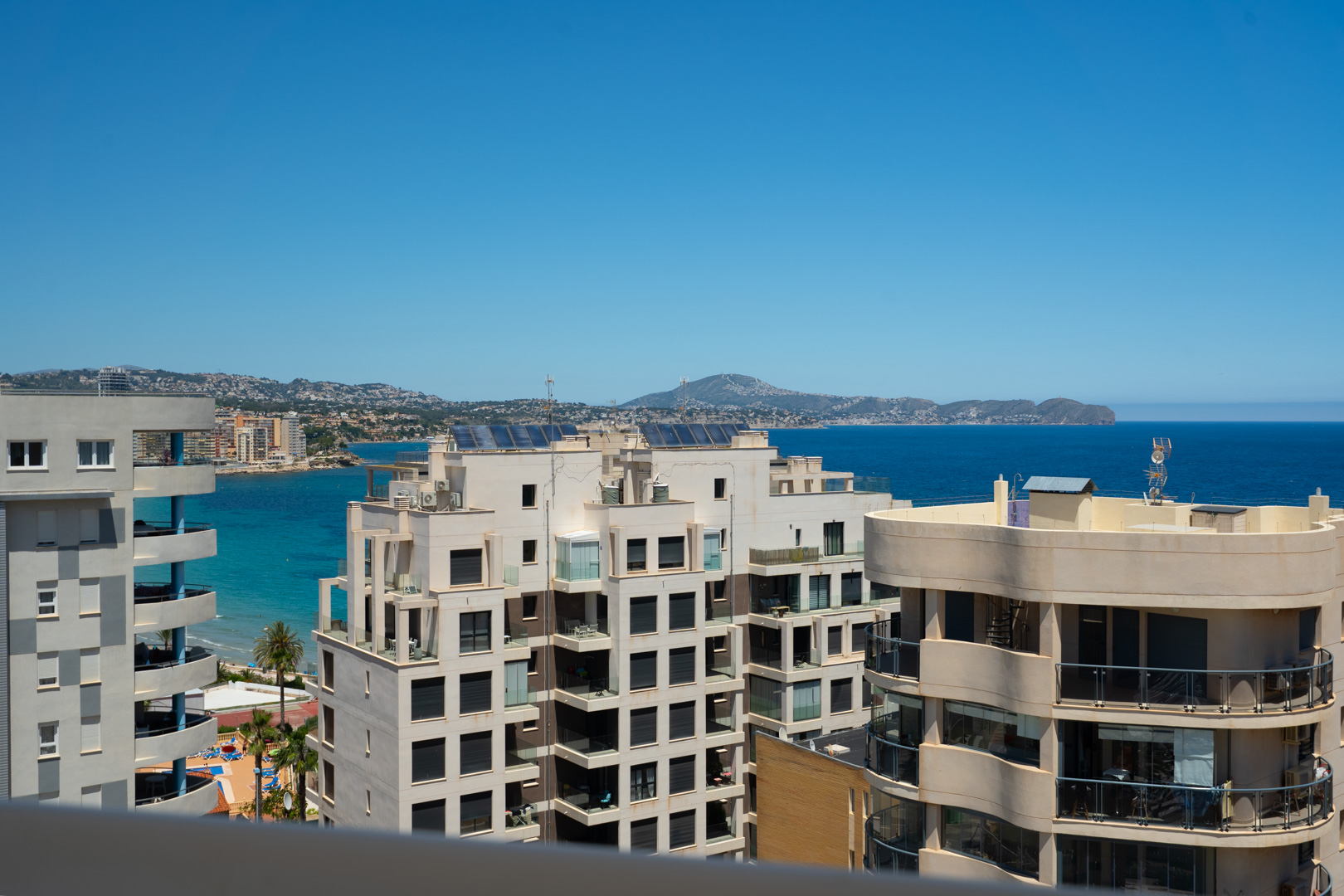 Panoramablick auf das Meer von einer Hochterrasse, mit moderner Küstenarchitektur, türkisfarbenem Wasser und fernen Bergen unter klarem blauem Himmel.