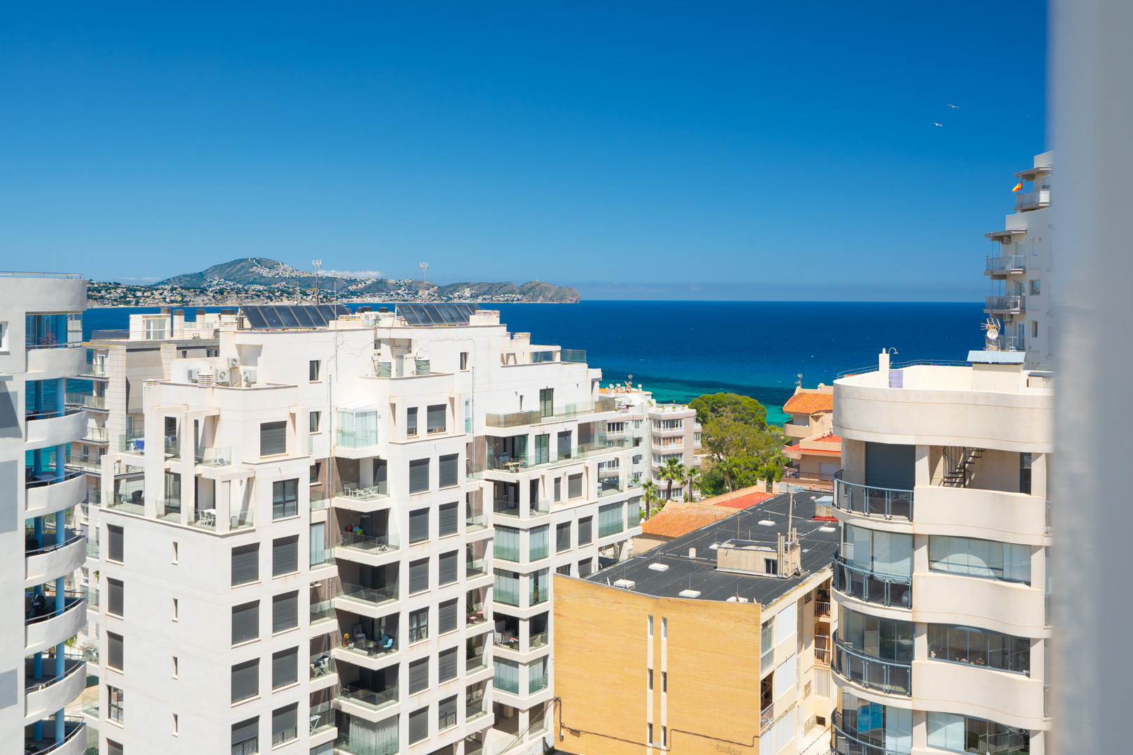 Panoramablick auf das Mittelmeer und die Küste von einer erhöhten Etage, mit Wohngebäuden im Vordergrund und fernen Bergen unter klarem blauem Himmel.