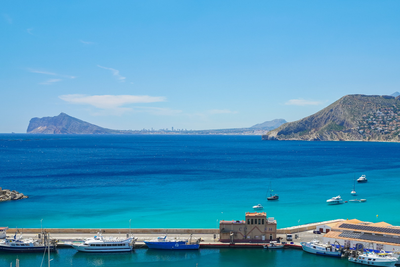 Panoramablick auf den Hafen von Calpe mit Booten, türkisfarbenem Wasser und dem ikonischen Peñón de Ifach im Hintergrund, umgeben von der Küstenbebauung.