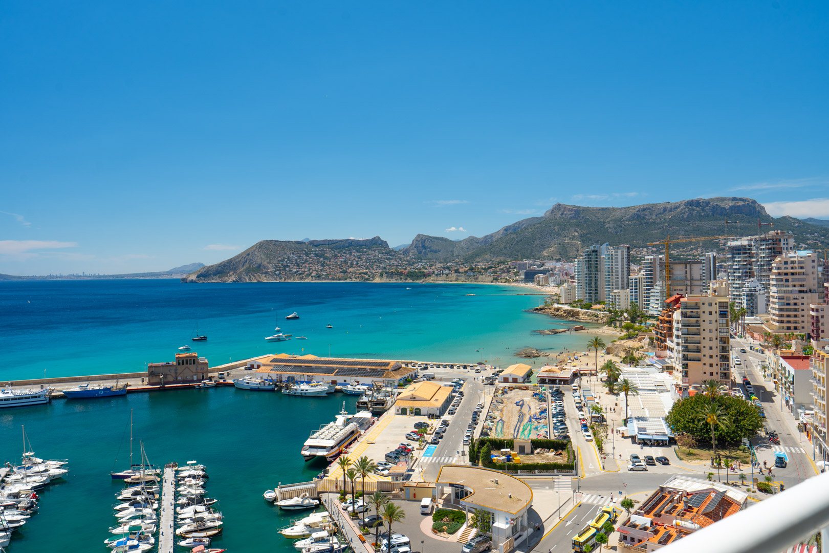 Panoramablick auf die Bucht von Calpe, den geschäftigen Yachthafen, Sandstrände und die Küstenstadt, umrahmt von fernen Bergen unter einem klaren blauen Himmel.