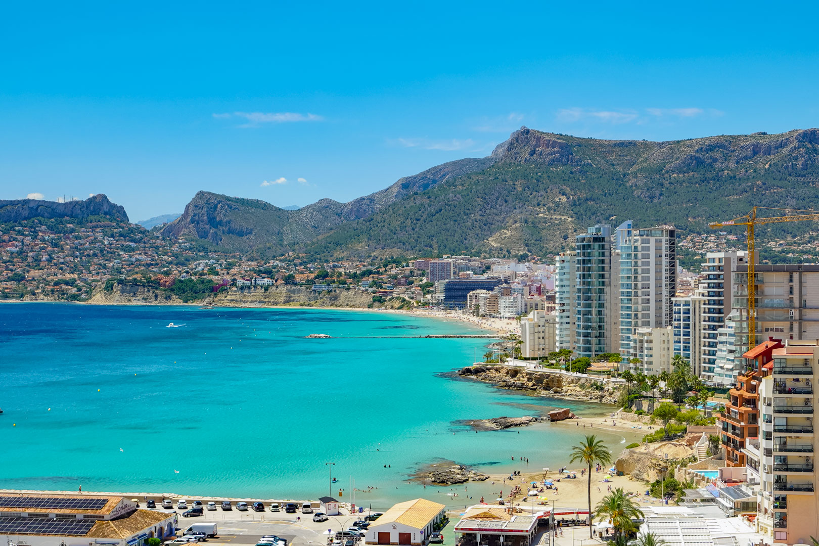 Panoramablick auf die Bucht von Calpe mit türkisfarbenem Wasser, Sandstränden, Küstengebäuden und fernen Bergen unter klarem blauem Himmel.
