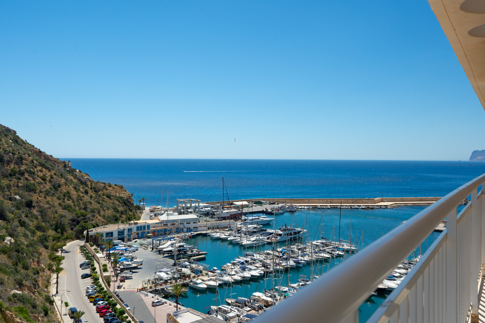 Panoramablick vom Balkon auf einen belebten Yachthafen mit zahlreichen Booten, das tiefblaue Meer und einen grünen Hügel unter klarem Himmel.