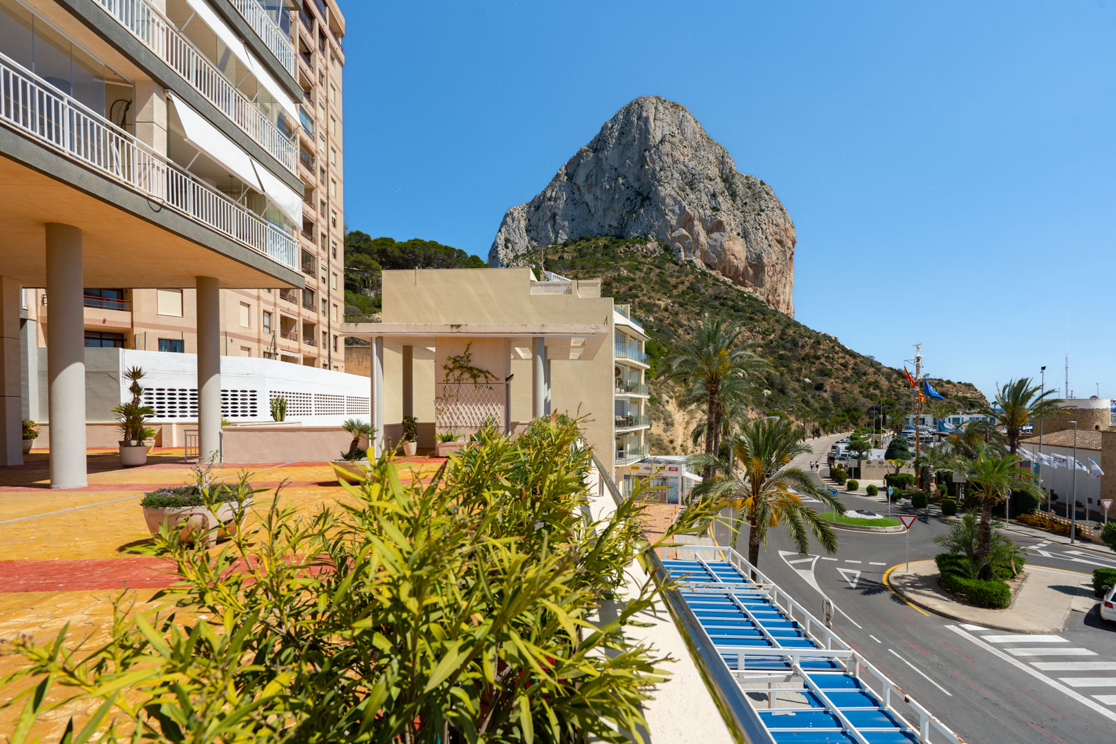 Panoramablick von einer Terrasse, mit dem ikonischen Peñón de Ifach, umliegenden Gebäuden und der Straße zum Hafen von Calpe unter klarem blauem Himmel.