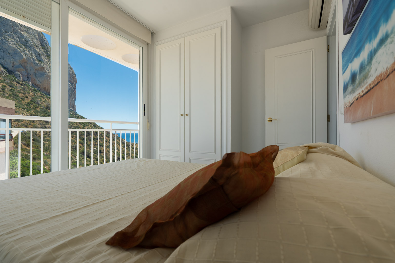 Schlafzimmer mit direktem Balkonzugang, mit Blick auf den markanten Peñón de Ifach, die grüne Hügellandschaft und das Mittelmeer. Ein weißer Einbauschrank ist vorhanden.
