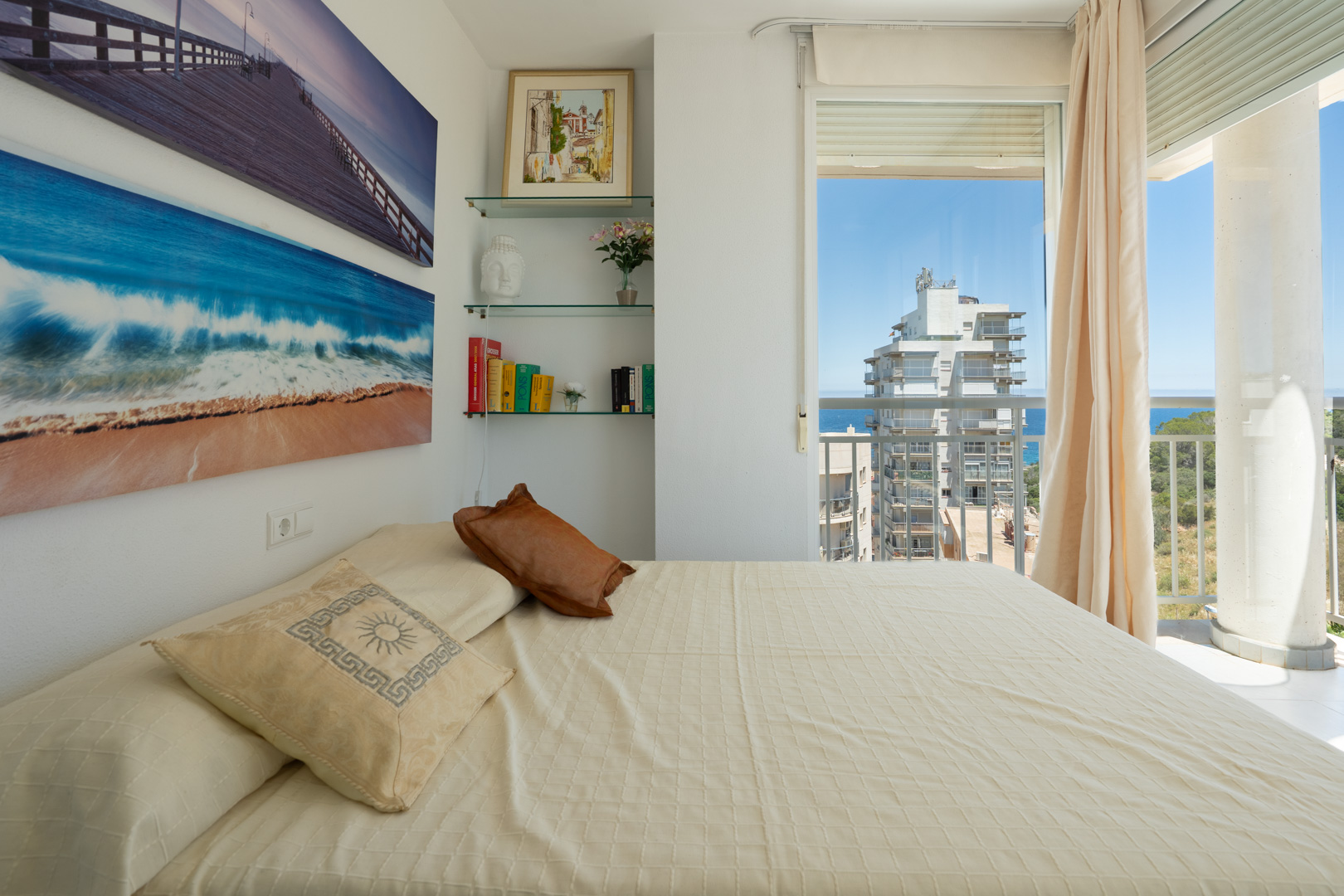 Schlafzimmer mit gemütlichem Bett, maritimem Wandschmuck und Glasregalen. Direkter Zugang zu einem Balkon mit teilweisem Meer- und Stadtblick.