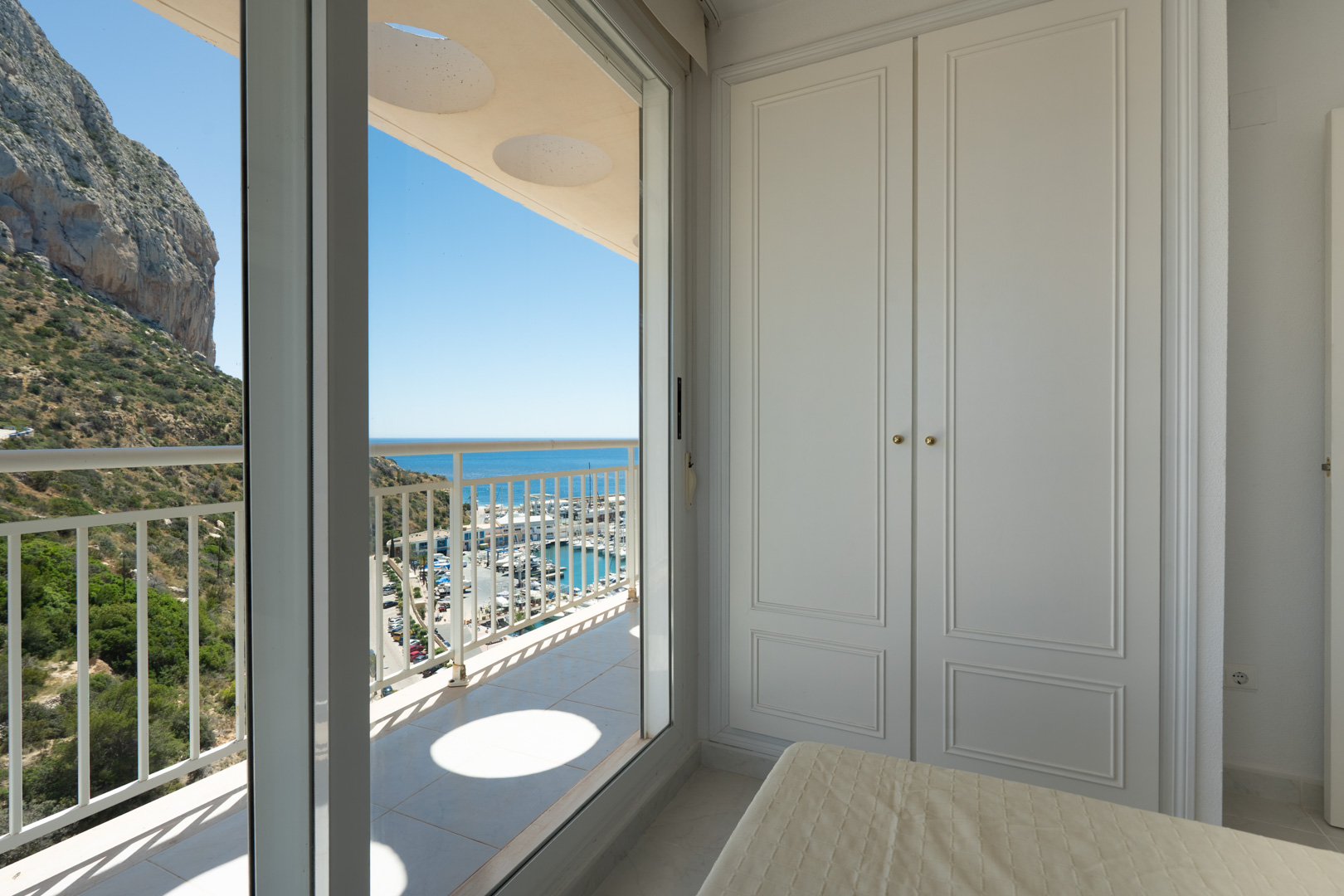 Schlafzimmer mit weißem Einbauschrank und Terrassenzugang. Panoramablick auf den Peñón de Ifach, den Yachthafen und das Mittelmeer unter blauem Himmel.