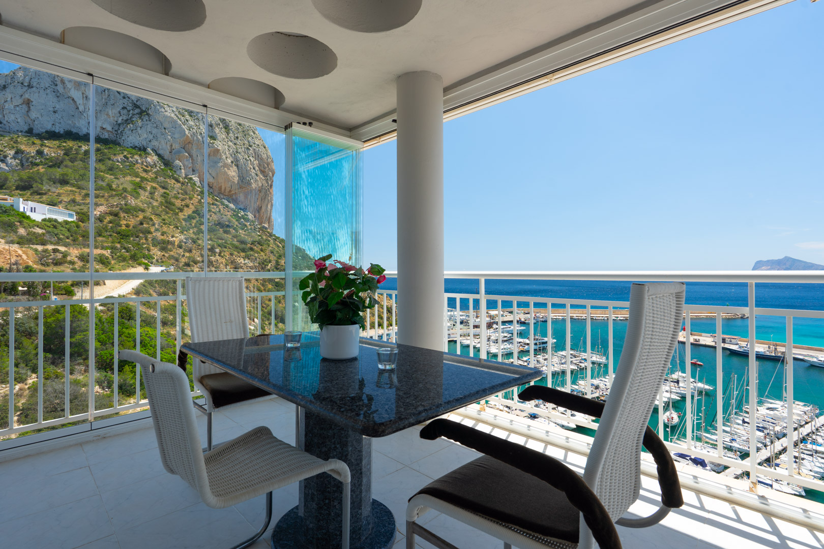 Überdachte Terrasse mit Granittisch und modernen Stühlen, ausgestattet mit Glasvorhängen, die einen Panoramablick auf den Peñón de Ifach und den Hafen von Calpe bietet.