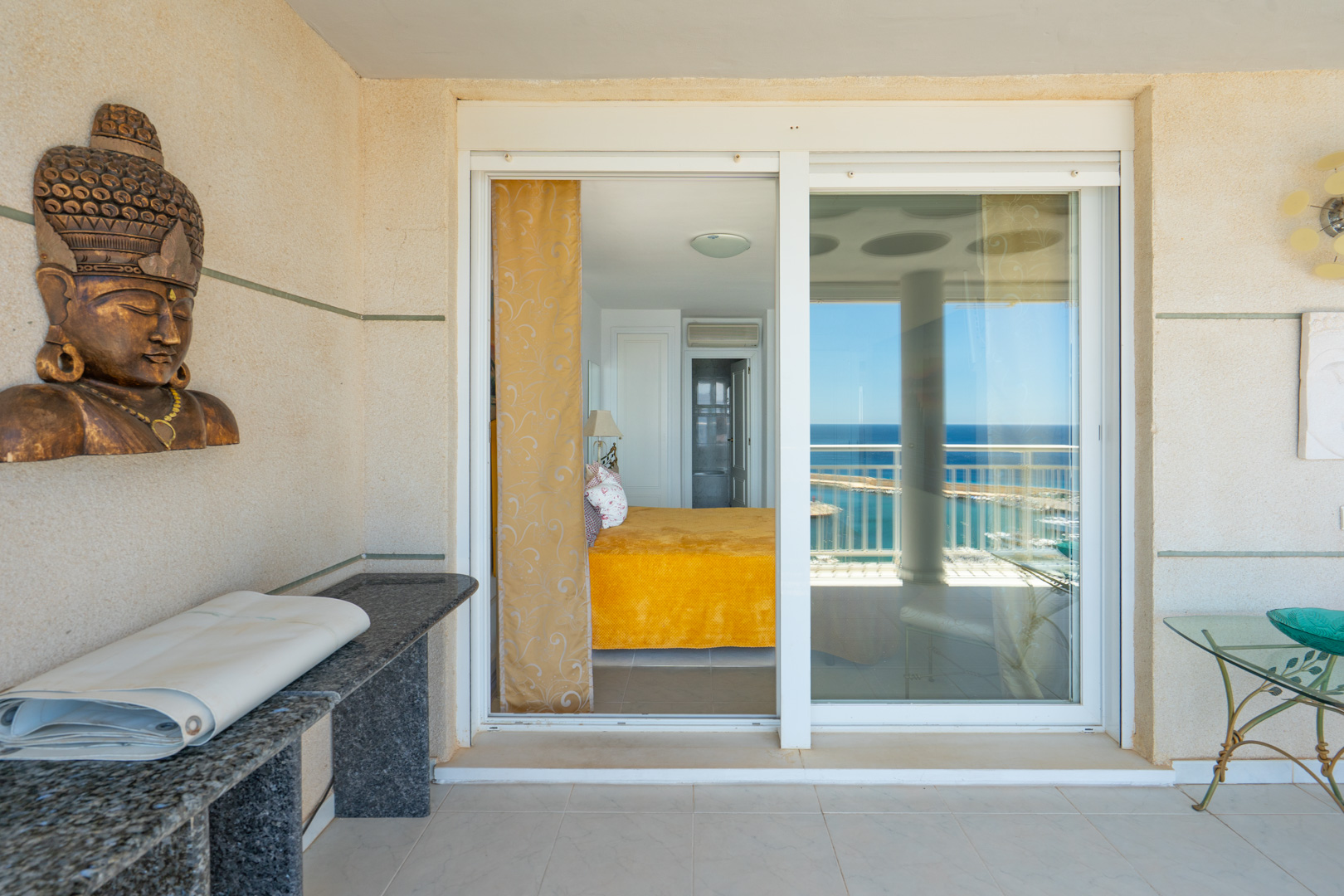 Überdachte Terrasse mit hellen Fliesen und strukturierten Wänden, mit Glasschiebetür zu einem Schlafzimmer, von dem aus Meerblick sichtbar ist. Eine Granitkonsole und Deko sind ebenfalls zu sehen.