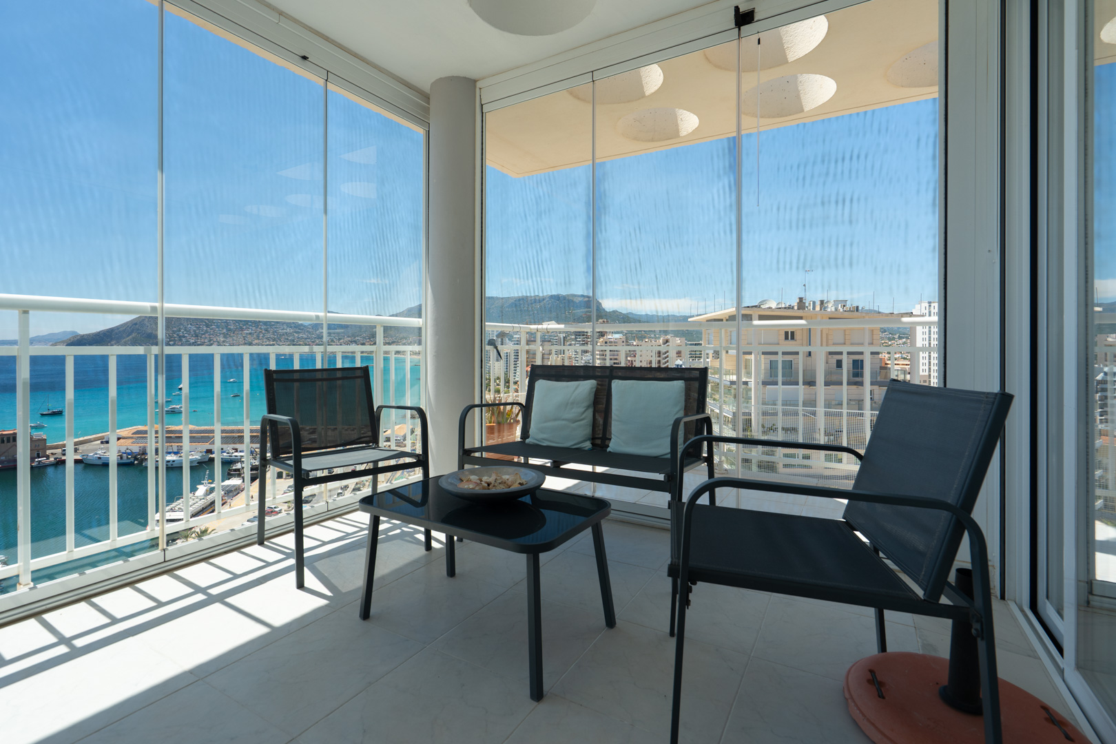 Überdachte Terrasse mit verschiebbaren Glasvorhängen, Outdoor-Möbeln und Panoramablick auf den Hafen von Calpe, das Meer und die Berge. Heller Fliesenboden.