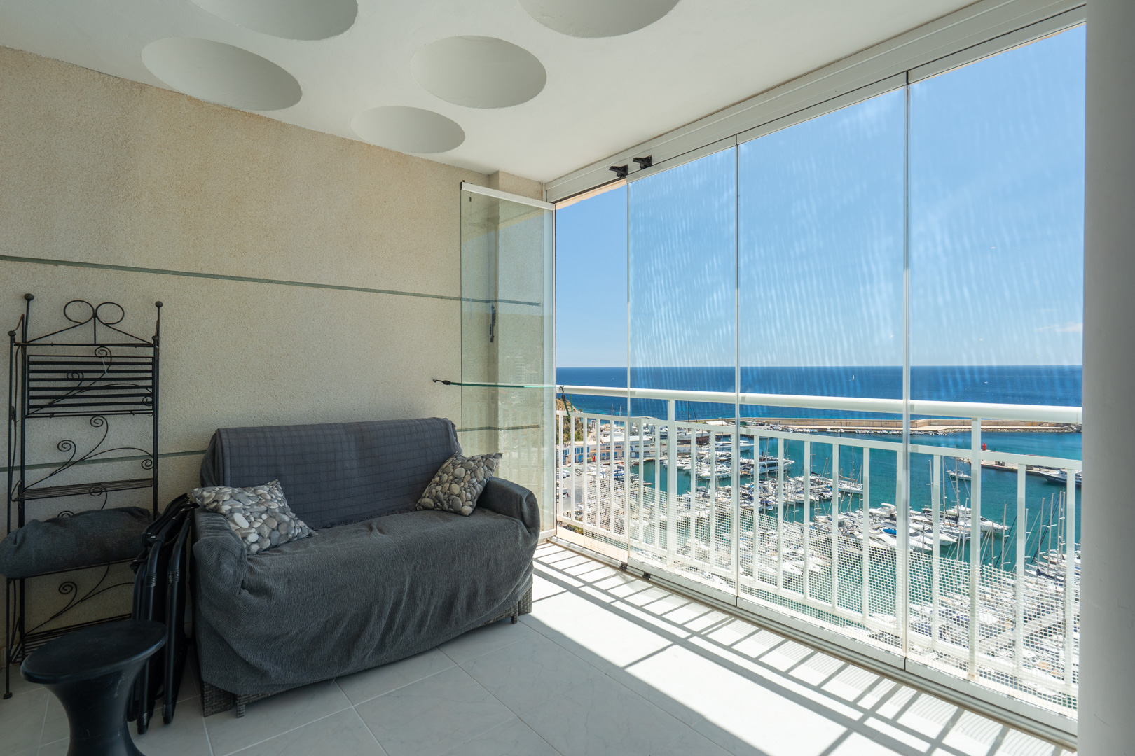 Verglaste Terrasse mit direktem Blick auf den Yachthafen und das Meer. Ausgestattet mit Sofa und schmiedeeisernem Regal, unter einer Decke mit runden Dekorelementen.