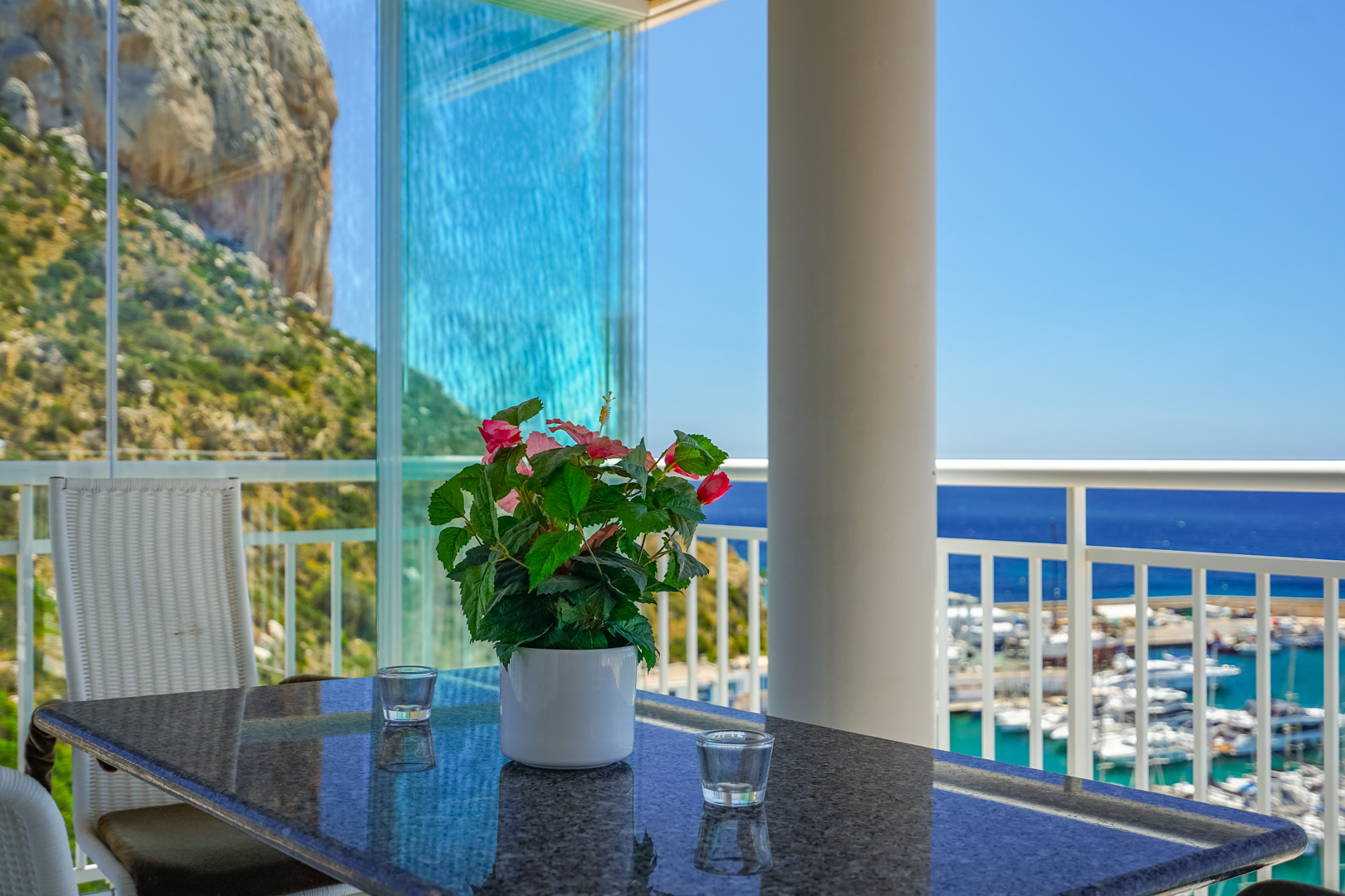 Verglaste Terrasse mit Glasvorhängen, direktem Blick auf den Peñón de Ifach und den Yachthafen von Calpe. Granittisch mit Blumenarrangement.