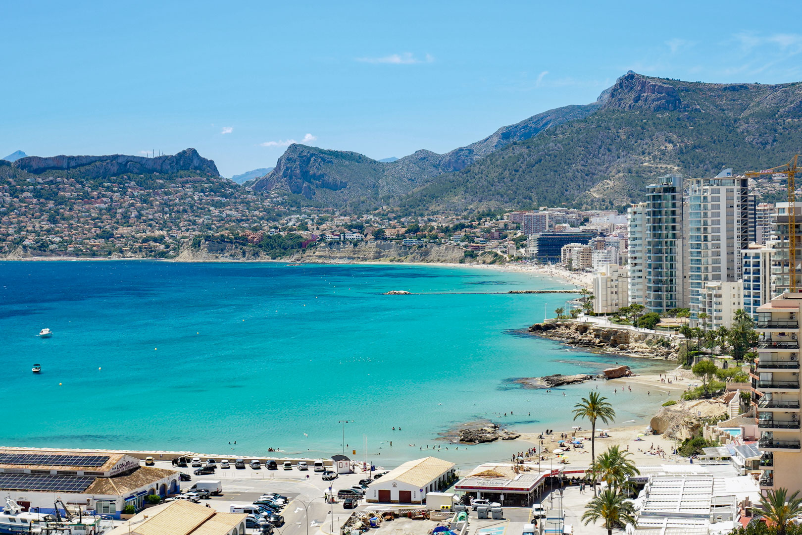 Weitläufiges Küstenpanorama der Bucht von Calpe, mit türkisfarbenem Wasser, Sandstränden, modernen Hochhäusern und fernen Bergen. Der Hafen mit Solarpaneelen ist im Vordergrund.