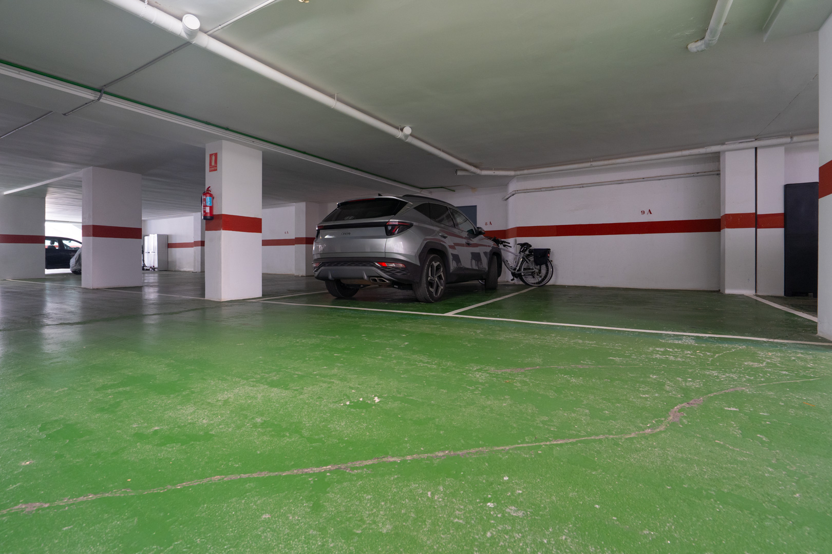 Zugewiesener Stellplatz in der Tiefgarage, mit grünem Betonboden und weißen Säulen mit roten Sicherheitsstreifen. Bietet Platz für ein Fahrzeug und ein Fahrrad.