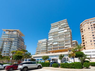 Außenansicht moderner Wohngebäude mit gestaffelten Glasbalkonen, eingebettet in eine sonnige Stadtlandschaft mit parkenden Autos und Palmen.