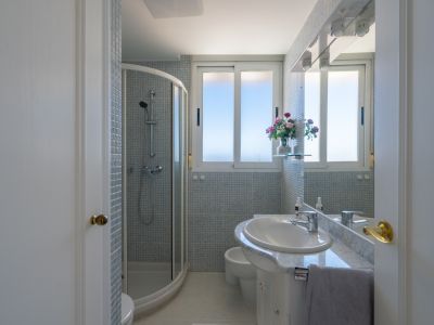 Badezimmer mit Eckduschkabine, weißem Waschtisch mit Marmorplatte und hellblauen Mosaikfliesen. Fenster für Tageslicht und beleuchteter Spiegel.