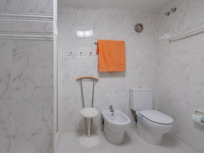 Badezimmer mit weißen Marmor-Effekt-Fliesen, ausgestattet mit WC, Bidet und einem praktischen Duschbereich mit Haltegriff. Eine Wanduhr und Handtuchhaken sind ebenfalls vorhanden.