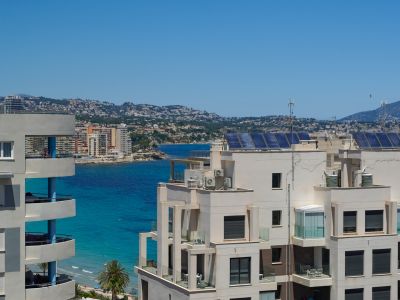 Blick von einer oberen Etage auf moderne Küstenarchitektur, mit verglasten Balkonen und Solarpaneelen auf den Dächern, umrahmt vom türkisfarbenen Meer und fernen Hügeln.