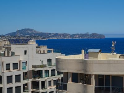 Erhöhter Blick über Dächer mit Solarpaneelen, das tiefblaue Meer und eine ferne Küstengebirgskette unter klarem Himmel.