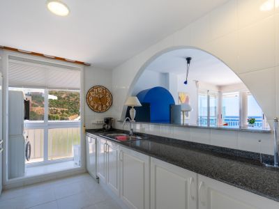 Funktionale Küche mit weißen Schränken, dunkler Granitarbeitsplatte und Waschküche am Fenster mit Blick auf den Hügel. Ein Bogen verbindet sie mit dem Wohnbereich und Meerblick.
