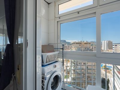 Funktionale Waschecke mit Waschmaschine und Wäschekorb, umgeben von Fliesenwänden und großen Fenstern, die einen urbanen Blick auf Gebäude, ferne Berge und einen Pool bieten.