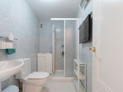 Funktionales Badezimmer mit hellblauen Mosaikfliesen an den Wänden, ausgestattet mit WC, Standwaschbecken und Duschkabine. Ein kleiner Unterschrank und zusätzliche Regale bieten Stauraum.