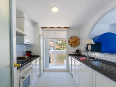 Galeerenküche mit weißen Schränken, dunklen Granitarbeitsplatten und Fliesenrückwand. Backofen, Kühlschrank und Waschmaschine im angrenzenden Hauswirtschaftsraum, mit Hügelblick.