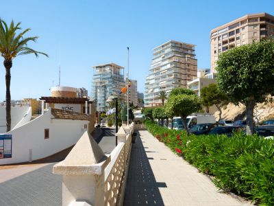 Gepflasterter Fußgängerweg mit Steinbalustrade und üppigen Oleanderbüschen, der auf moderne Hochhäuser und den Eingang des Real Club Náutico Calpe unter klarem blauem Himmel zuführt.