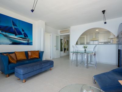 Geräumiger Wohn-Essbereich mit blauem Sofa, Glastisch und transparenten Stühlen, akzentuiert durch ein großes Segelboot-Kunstwerk. Ein Bogen führt zur modernen, halboffenen Küche.