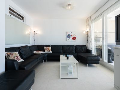 Geräumiges Wohnzimmer mit großem schwarzen Ecksofa und Zierkissen, weißen Fliesenboden und modernem weißen Couchtisch. Glasschiebetüren führen direkt zur Terrasse.