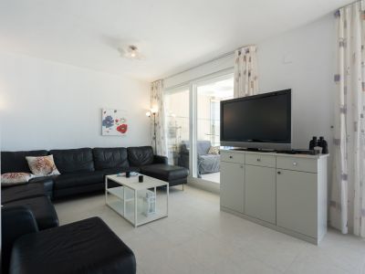 Geräumiges Wohnzimmer mit schwarzem Lederecksofa, weißem Couchtisch und Fliesenboden. Glasschiebetüren führen zu einer Terrasse, ergänzt durch einen TV-Schrank und gemusterte Vorhänge.