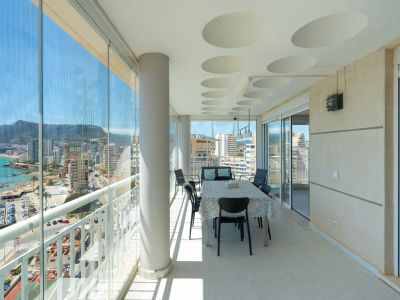 Großzügige verglaste Terrasse mit Essbereich und Panoramablick auf Meer, Hafen und Berge von Calpe. Sie verfügt über Glasvorhänge, eine Decke mit runden Akzenten und einen gefliesten Boden.