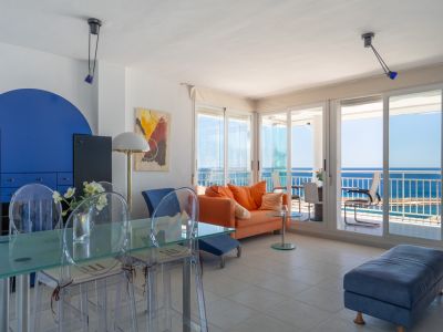 Großzügiger Wohn-Essbereich mit orangefarbenem Sofa, Glastisch und transparenten Stühlen, akzentuiert durch eine blaue Wand. Große Schiebetüren führen zu einer Terrasse mit direktem Meerblick.