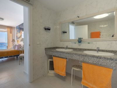 Hauptbadezimmer mit Doppelwaschtisch aus Granit, Marmorwänden und verziertem Spiegel, mit direktem Blick ins Meerblick-Schlafzimmer.