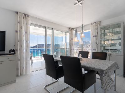 Heller Essbereich mit modernem Tisch und Stühlen, direkter Zugang zur verglasten Terrasse mit Panoramablick auf Meer und Peñón de Ifach. Vitrine mit Geschirr.