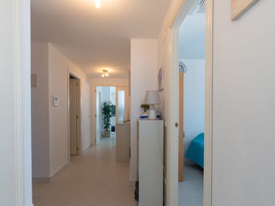 Heller Flur mit hellen Fliesen, weißen Türen und einer Konsole mit Lampe. Ein Schlafzimmer mit blauem Bett und ein weiterer Raum mit Pflanze sind sichtbar.