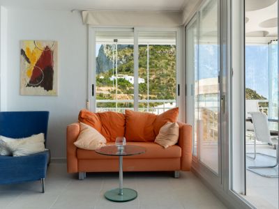 Heller Wohnbereich mit orangefarbenem Sofa und blauem Sessel. Große Schiebetüren führen zu einer Terrasse mit Bergblick, Außenmöbeln und einer Decke mit markantem Kreisdesign.