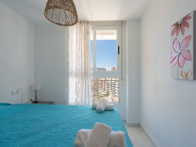 Helles Schlafzimmer mit blauem Steppbett und weißen Handtüchern. Das Fenster bietet Stadt- und Bergblick, ergänzt durch eine geflochtene Hängelampe und ein Blumenbild.