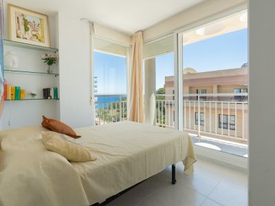 Helles Schlafzimmer mit Doppelbett, dekorativen Glasregalen und direktem Zugang zur Terrasse mit teilweisem Meerblick und Stadtansicht.